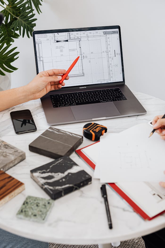 Interior designers reviewing tile samples and floor plans using a laptop and pen on a marble table.